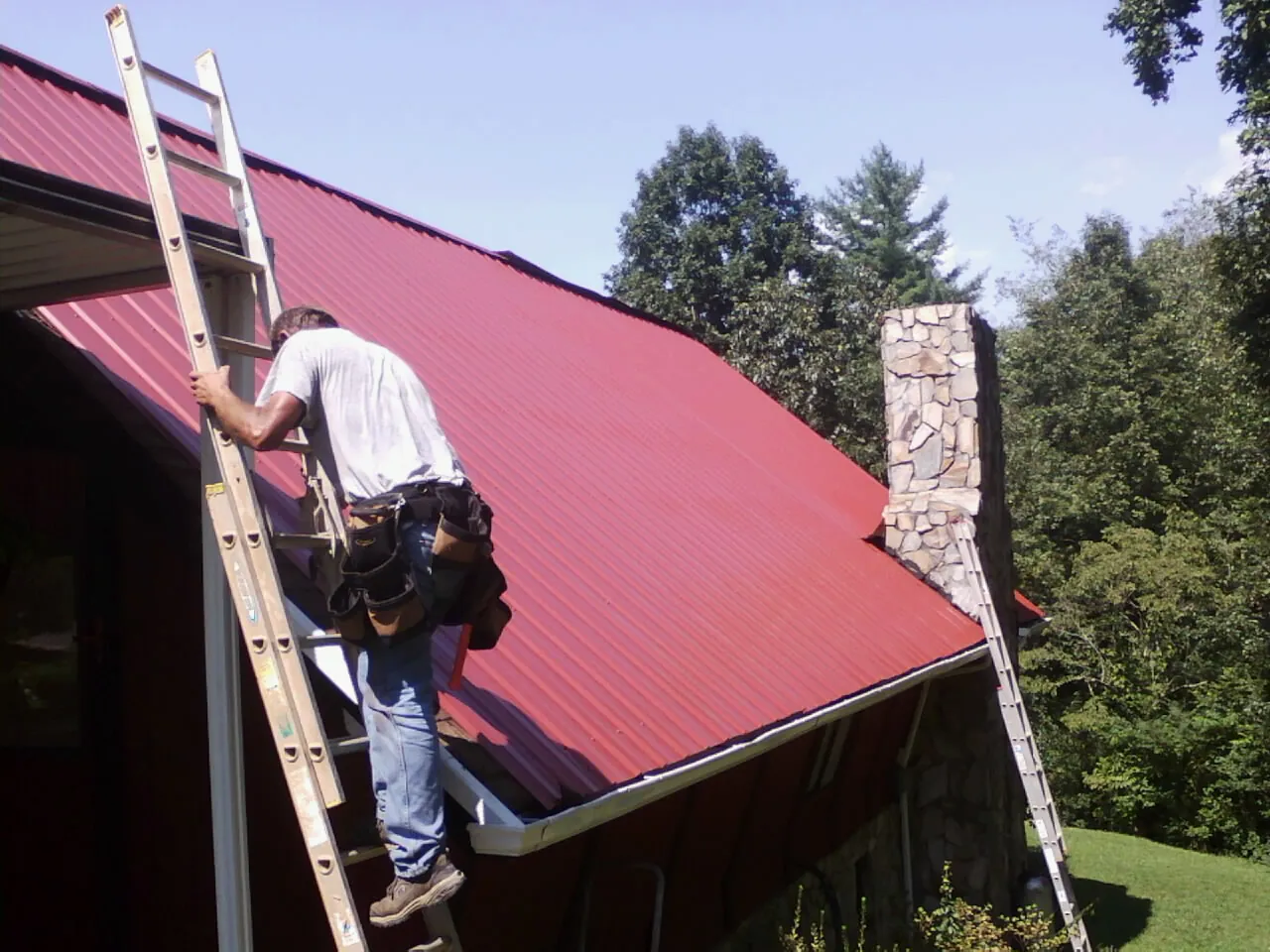 Roofer on a ladder beside a red metal roof and stone chimney — Soffit & Fascia Repair by Roofing Brothers in Slatington